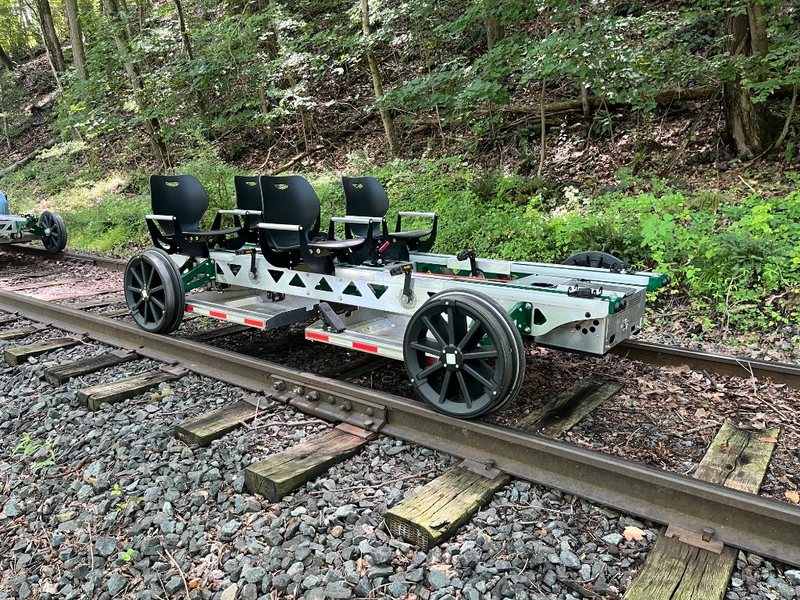 A green four-seat Rail Glider on the rails in the woods, showing adjustable seats, a plastic basket, and a metal floor under the passengers.